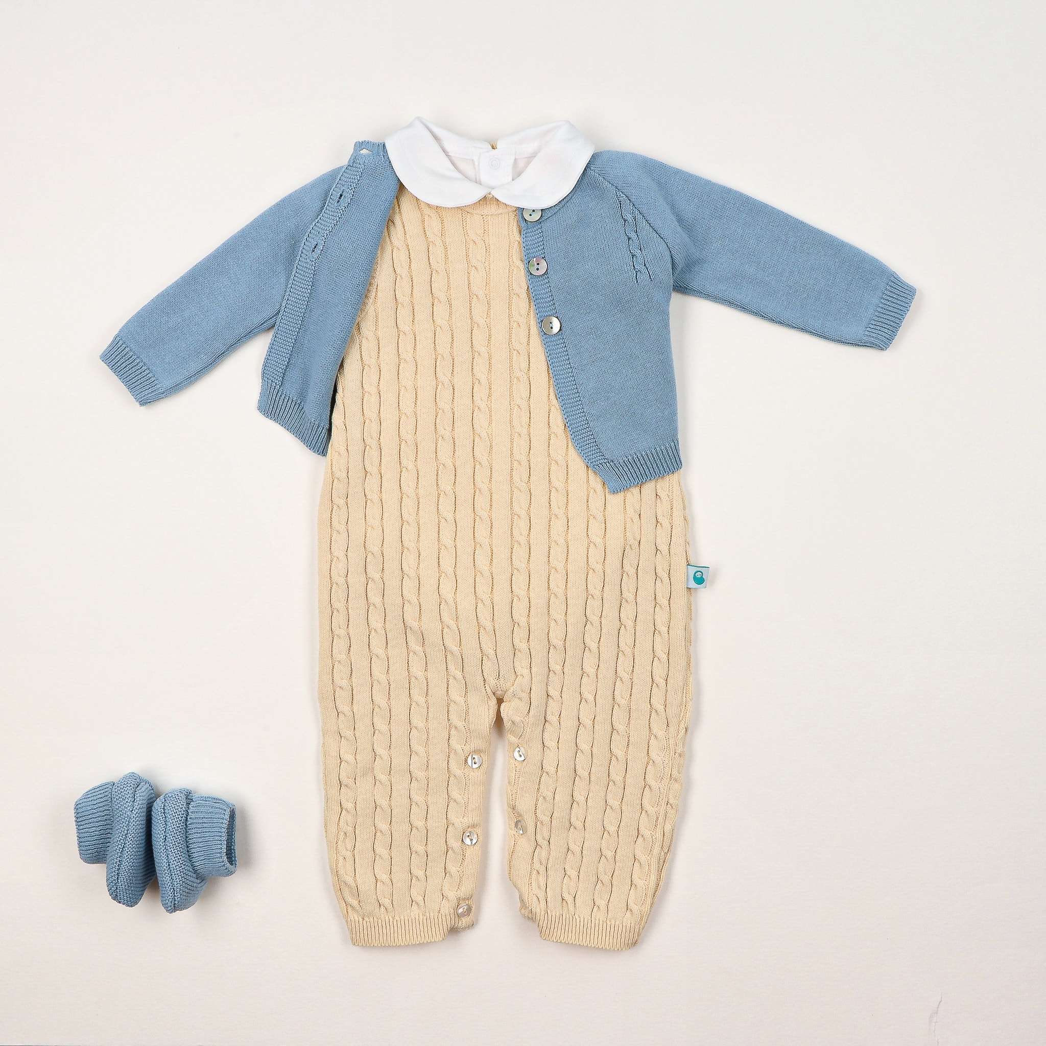 Look completo composto por jardineiras amarelas tricotadas em ponto trança, com botões madrepérola no entrepernas; casaco tricotado azul inglês com detalhes de ponto trança no peito e botões madrepérola na frente; body gola redonda com bordado espiga em branco; carapins tricotados azul inglês.