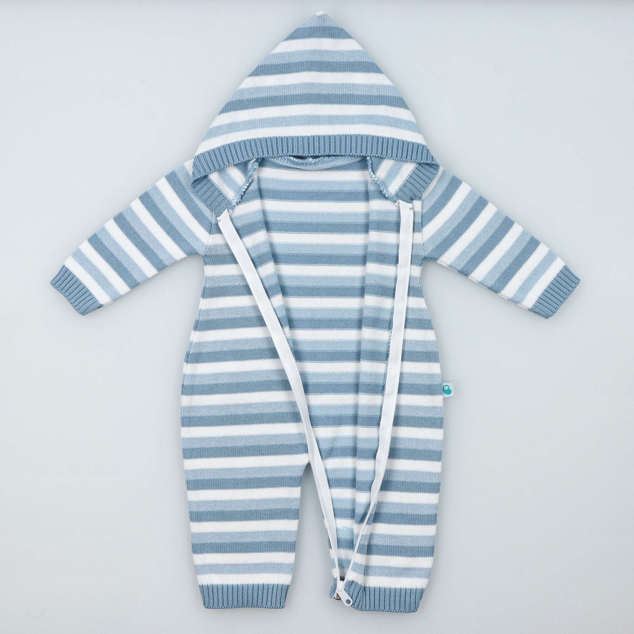 Vista de frente, sobre fundo cinzento, de macacão em malha tricotada 100% algodão, com carapuço e fecho de correr em plástico aberto até ao pezinho. Padrão de riscas brancas, azul claro e azul escuro.