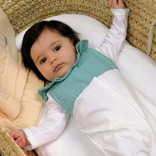 Bebé deitado numa alcofa de vime vestido com um babygrow branco com o peito, gola e pés em tecido plumeti verde. Tem uma manta amarela ao lado.