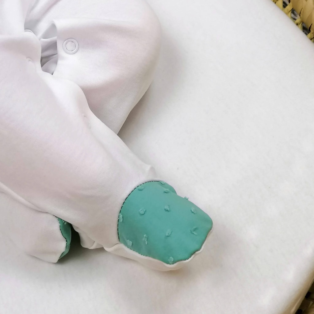Pormenor do pé de um bebé deitado numa alcofa em tecido plumeti verde de um babygrow para bebé branco.