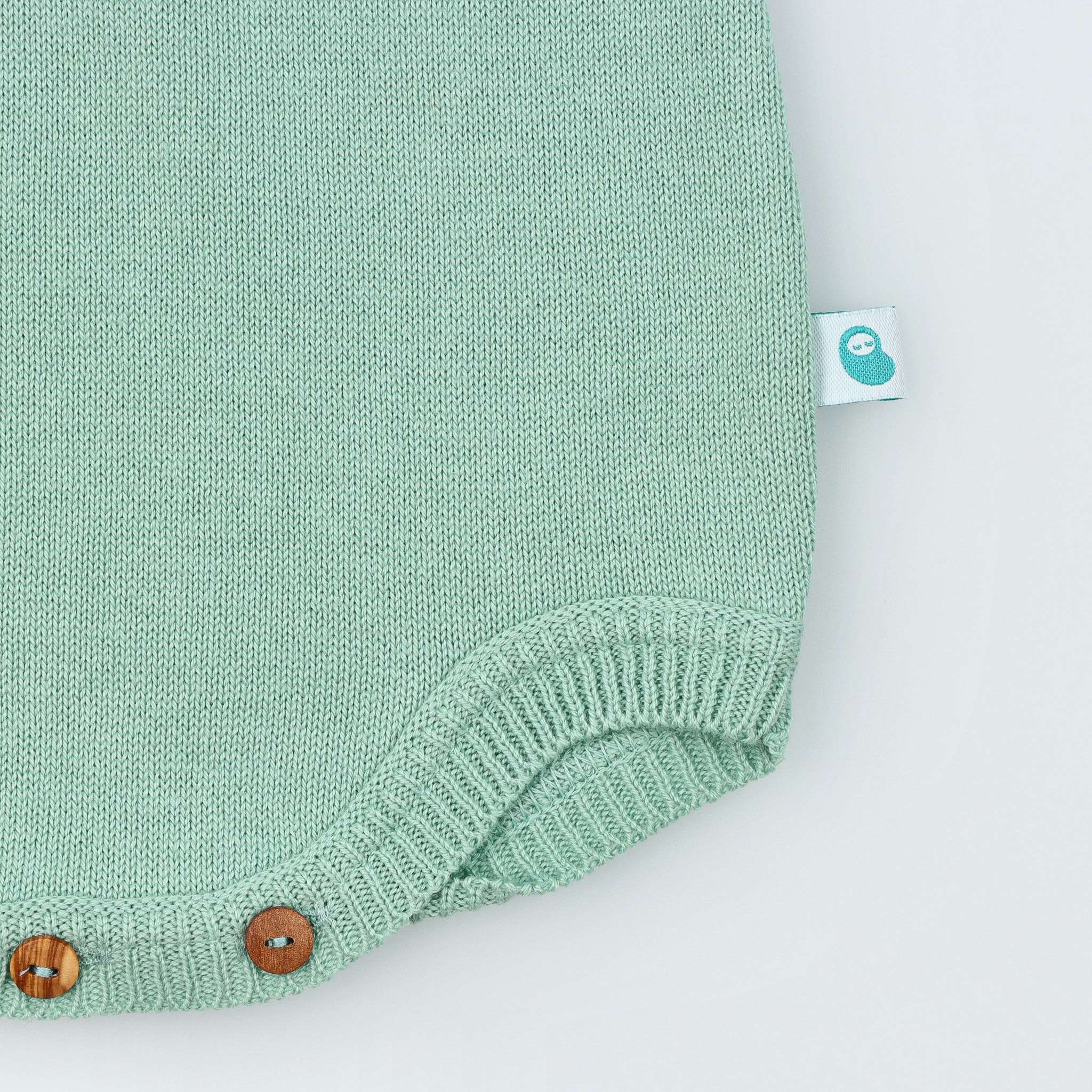 Detalhe de etiqueta Bean do lado esquerdo do fofo de bebé em malha tricotada 100% algodão verde seco. Vista de frente e fundo cinzento.