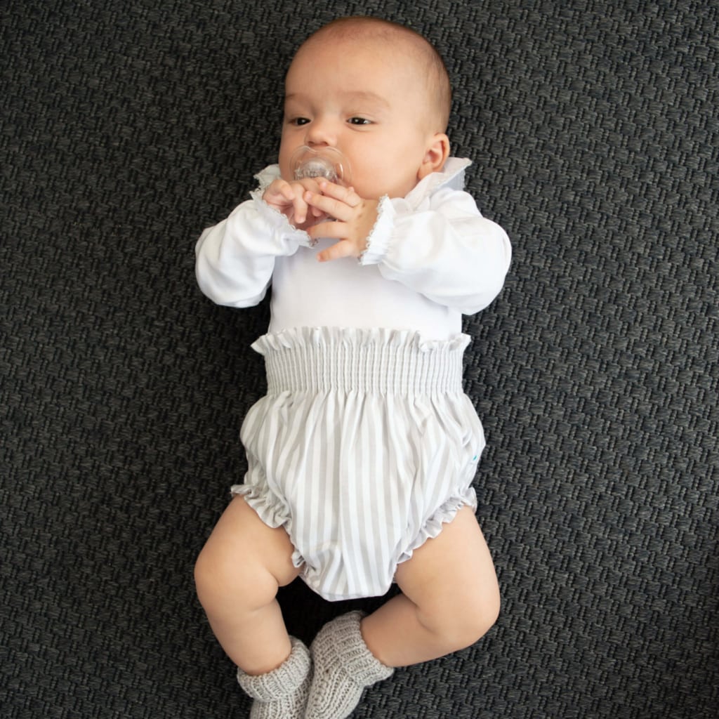Bebé com body branco e tapa fraldas cinzento.